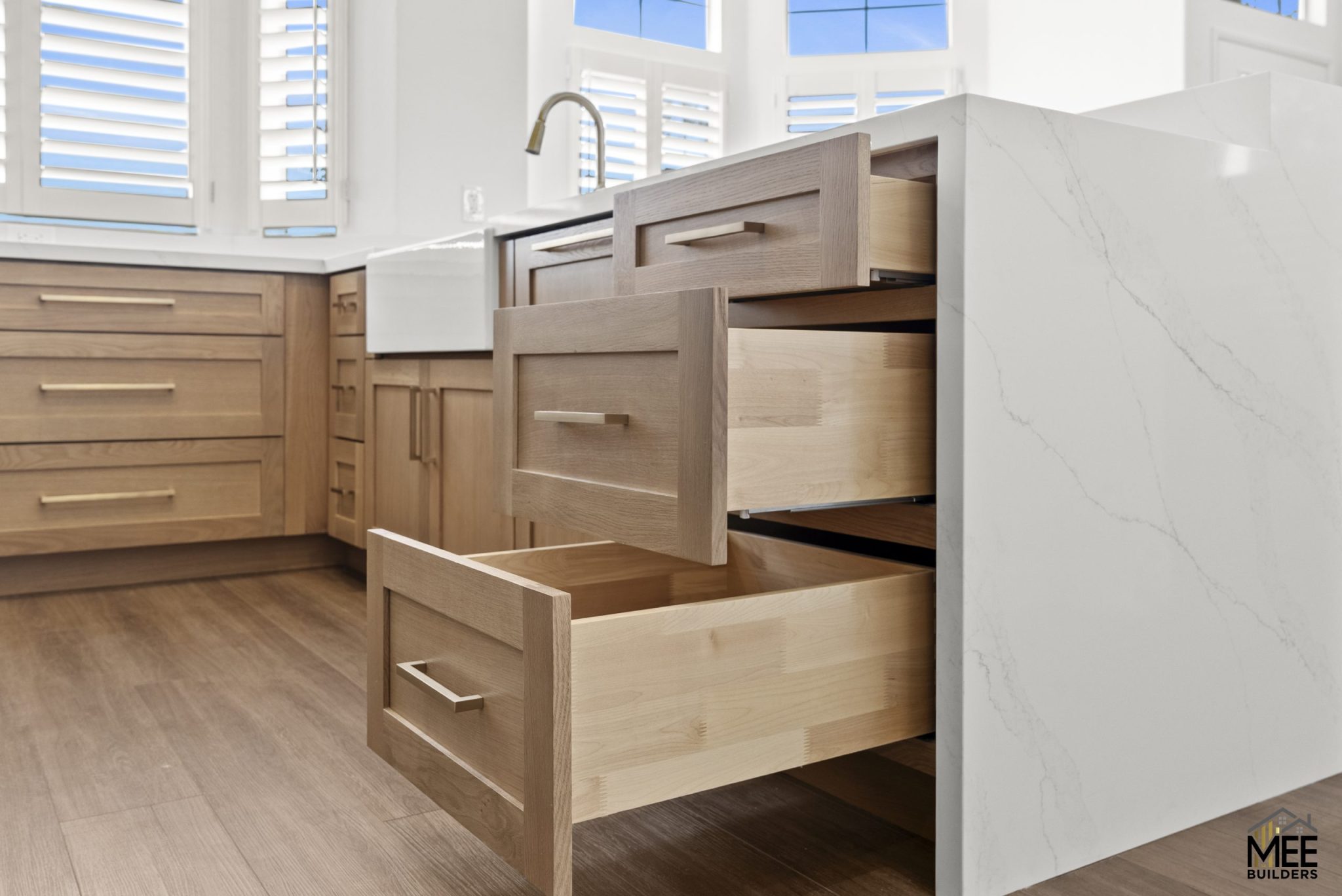 A modern kitchen featuring multiple pull-out drawers with light wood construction and sleek metal handles, showcasing custom cabinet storage solutions. The drawers are extended to display their deep interior space and dovetail joinery, set against white marble-look countertops and matching natural wood cabinetry. Large windows with white shutters provide natural lighting to the bright, contemporary kitchen space.