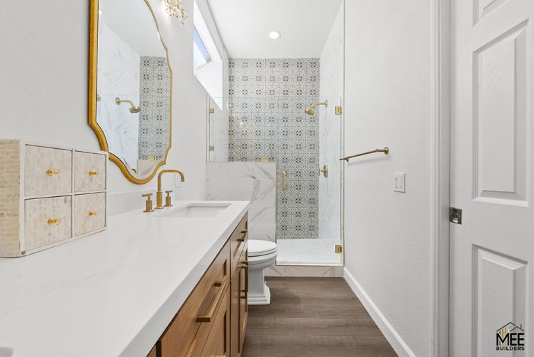 A modern bathroom featuring a white floating vanity with natural wood drawers, gold fixtures throughout, and decorative patterned tile in the shower area. The space includes a gold-framed mirror, white walls, dark wood flooring, and a glass-enclosed shower with a skylight above providing natural light.