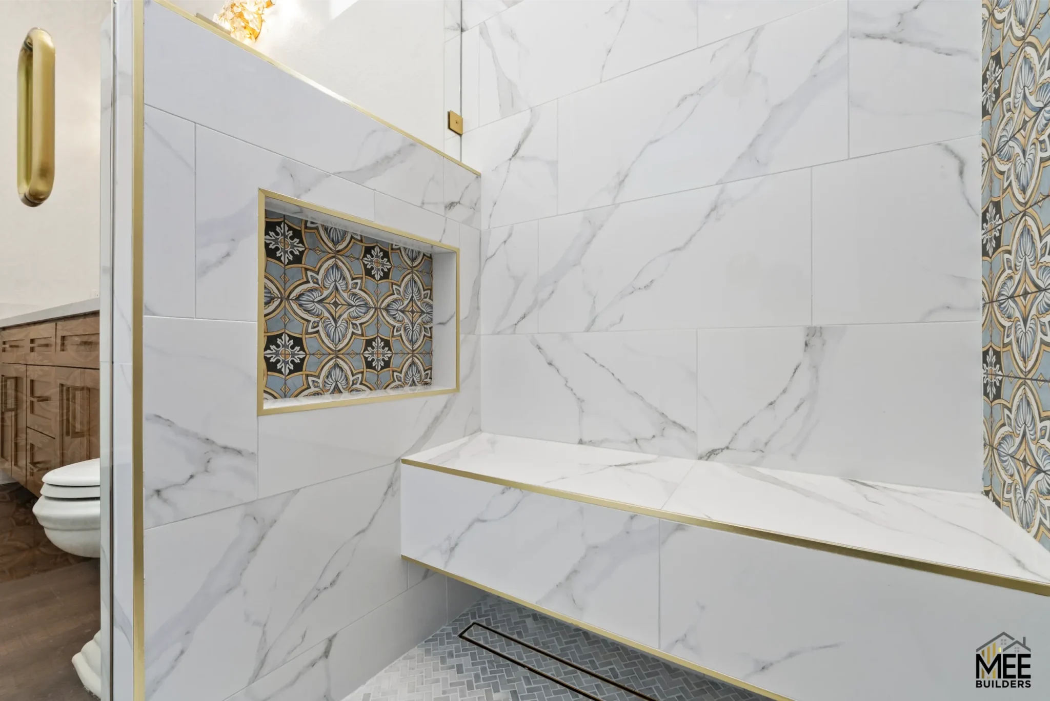 A luxury bathroom shower featuring white marble-look tiles with gray veining covering the walls and built-in bench. The space is accented with gold trim along the edges and includes a decorative niche with ornate blue and gold patterned tiles. The floor displays a herringbone mosaic pattern, and the shower includes a glass door with gold hardware.
