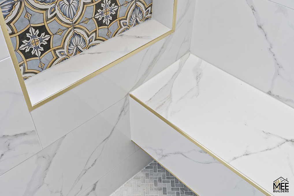 A downward view of elegant white marble stairs with gold metal trim accents along the edges. The steps feature sophisticated veining patterns in the marble, while decorative ornate tilework with geometric and floral motifs in blue, gold, and black is visible at the top landing. The Mee Builder logo appears in the bottom right corner of the architectural photograph.