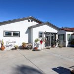 A modern white stucco house with dark gray trim features a distinctive geometric design with two peaked rooflines and rectangular windows. The front entrance is decorated with holiday wreaths and potted plants, while the wide concrete driveway leads to an attached garage visible on the right. Desert landscaping with cacti and decorative garden elements adorns the front yard under a clear blue sky.