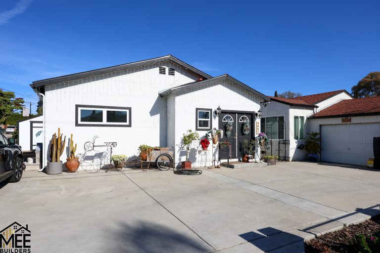 A white stucco single-story home with a spacious concrete driveway, featuring black-trimmed windows and decorative wreath-adorned double doors at the entrance. The front yard displays various potted plants, cacti, and decorative elements including a wire bicycle sculpture, set against a clear blue sky. The property is flanked by neighboring homes with a garage visible on the right side.