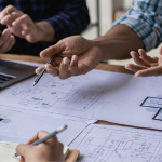 A collaborative construction or architecture team meeting around a table covered with blueprints and floor plans. Multiple hands are visible pointing at and discussing the technical drawings, with one person holding a pen. A laptop, measuring tape, and yellow hard hat are visible on the workspace, indicating an active planning or design session.