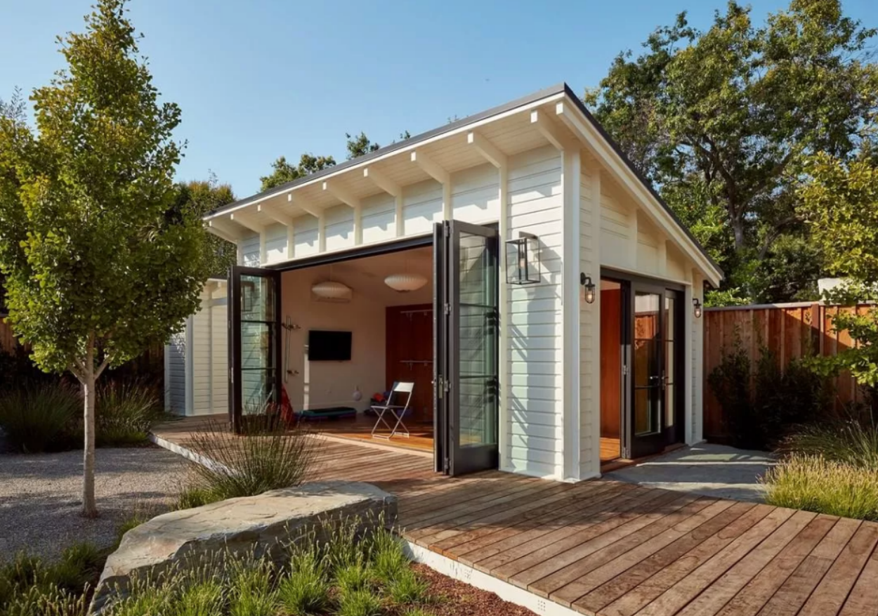 A modern backyard studio or accessory dwelling unit with white horizontal siding and a sloped roof featuring exposed beams. The structure has large black-framed bifold glass doors that open onto a wooden deck, revealing a minimalist interior with white walls and wood flooring. The building is surrounded by landscaping including young trees, ornamental grasses, and a wooden fence, set against a clear blue sky.