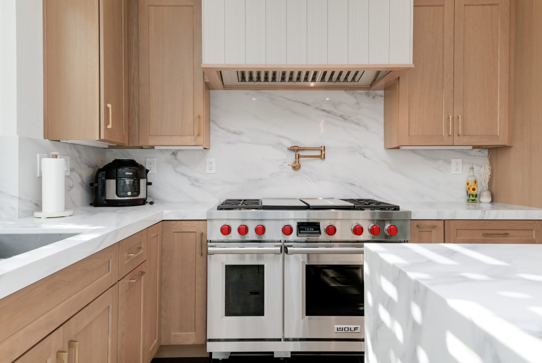 A modern kitchen featuring light wood shaker-style cabinets and white marble countertops with gray veining. The centerpiece is a professional Wolf dual-oven range with distinctive red control knobs, flanked by a gas cooktop. The backsplash continues the marble theme, with a brass pot filler mounted above the range and a stainless steel hood vent beneath white upper cabinets.