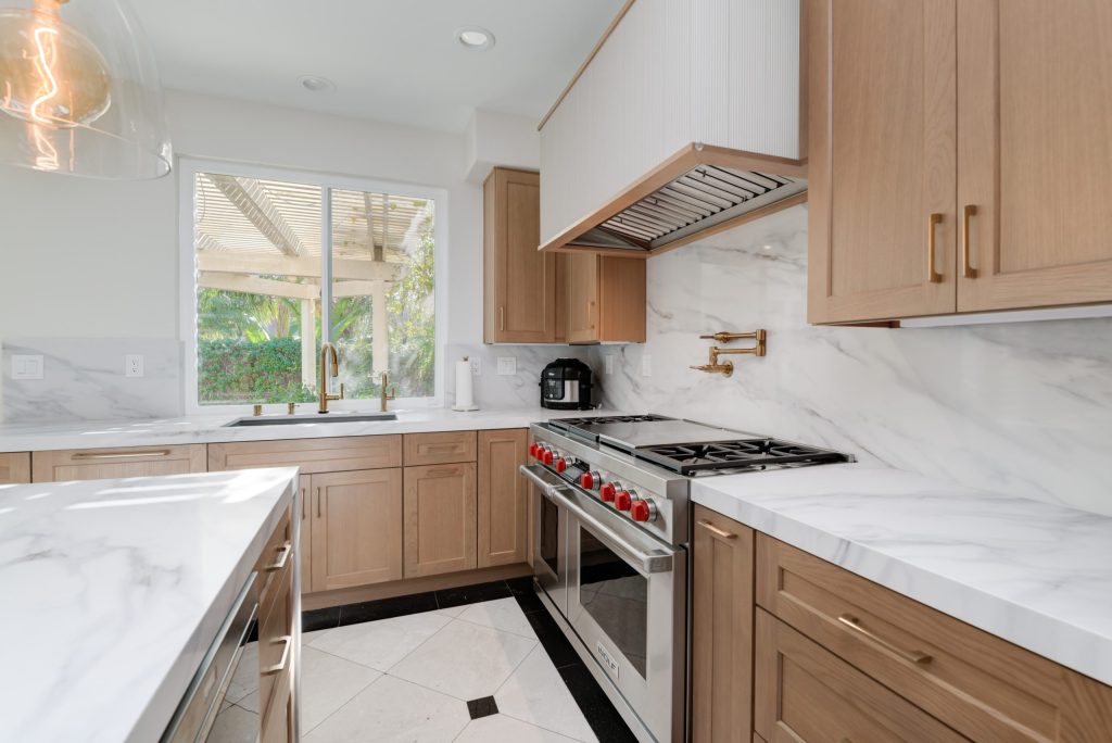 A modern kitchen featuring light wood cabinetry and white marble countertops and backsplash. The space includes a professional-grade gas range with red control knobs, stainless steel appliances, and a custom range hood. Natural light streams through large windows overlooking a covered patio area, while the room is anchored by light tile flooring and contemporary fixtures including brass hardware and pot filler.