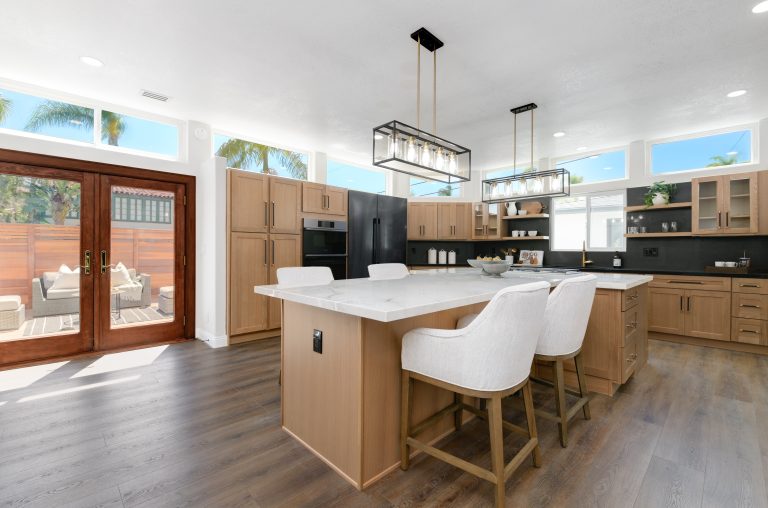 A modern open-concept kitchen featuring light wood cabinetry, a large white marble island with four white upholstered bar stools, and black industrial-style pendant lighting. The space showcases French doors leading to an outdoor patio, high transom windows allowing natural light, and a mix of black and stainless steel appliances against a dark backsplash. The room has gray-toned hardwood flooring and a bright, airy atmosphere with views of palm trees outside.