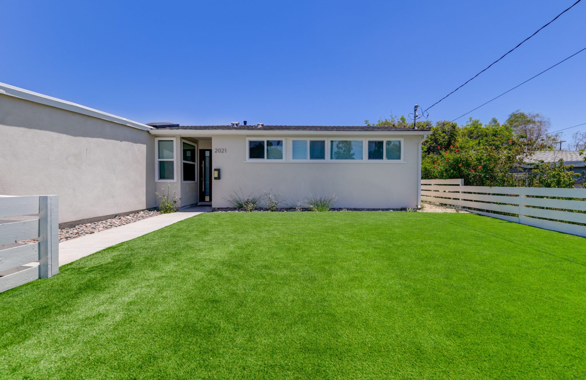 A single-story mid-century modern home with gray stucco exterior and flat roof, featuring a row of horizontal windows along the front facade. The house has a concrete walkway leading to the entrance, surrounded by a well-maintained green lawn with ornamental grasses and a white horizontal fence on the right side. The property is photographed under clear blue skies with mature trees visible in the background.