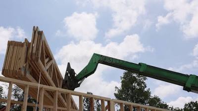 A green construction boom lift extends upward against a blue sky with white clouds, positioned near wooden building framing. The aerial lift appears to be in use at an active construction site, with exposed lumber framework visible in the foreground and trees in the background.