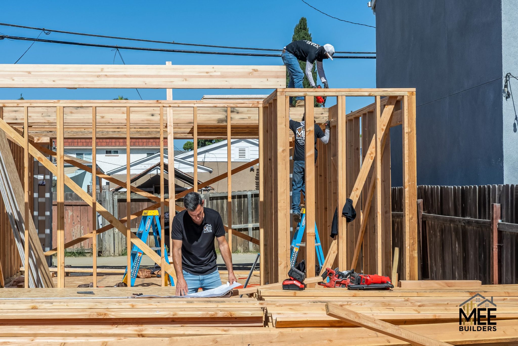 Construction workers frame a wooden structure on a bright sunny day. One worker stands on the floor working with materials while two others work on the top beam and wall frame. The setting shows a residential construction site with power lines, fencing, and neighboring buildings visible in the background, with the MEE Builders logo in the bottom right corner.