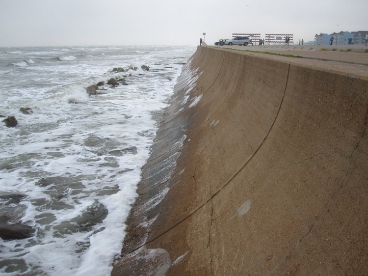 A curved concrete seawall rises steeply from a rocky shoreline where waves crash against the base. The protective barrier separates the turbulent ocean waters from a promenade and buildings visible in the background. A single person stands at the top of the wall, providing scale to the impressive coastal defense structure under an overcast sky.