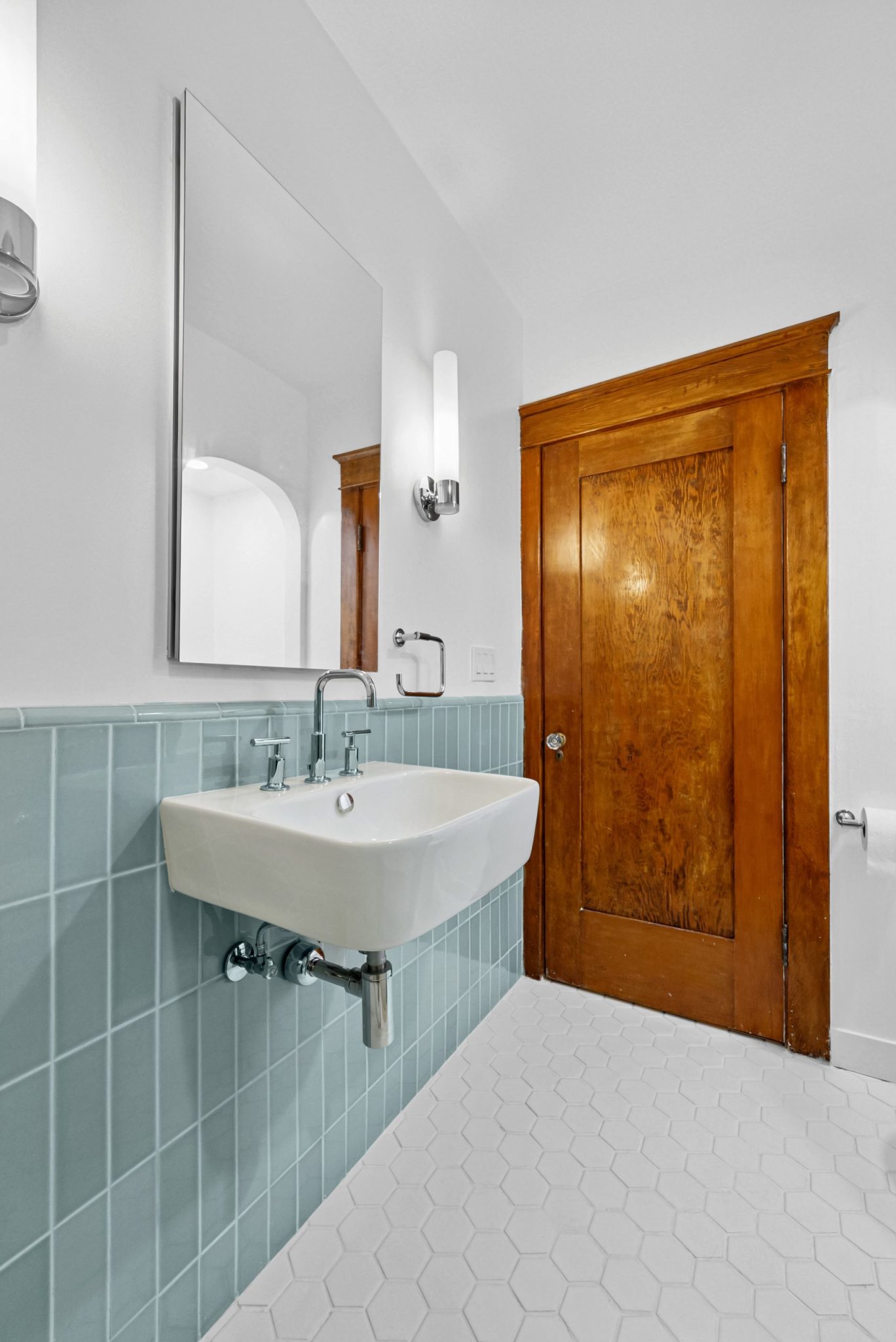 A bright bathroom featuring white hexagonal floor tiles and pale blue vertical subway tiles on the lower wall. A white wall-mounted sink with chrome fixtures sits beneath a large frameless mirror, while a wooden door with visible grain stands against the white upper walls. The space has vaulted ceilings and combines classic design elements with a clean, minimalist aesthetic.