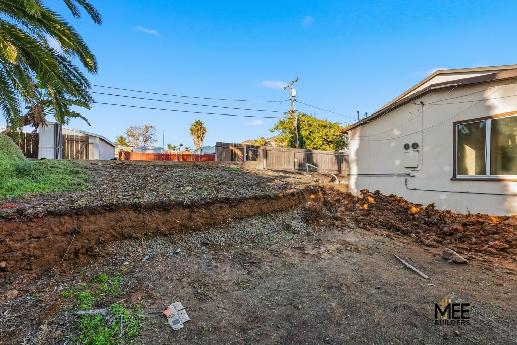 A residential construction site showing excavated and graded land in a backyard, with exposed soil layers and dirt piles. A single-story house with light-colored siding is visible on the right, while wooden fences, palm trees, and neighboring structures can be seen in the background under a clear blue sky.