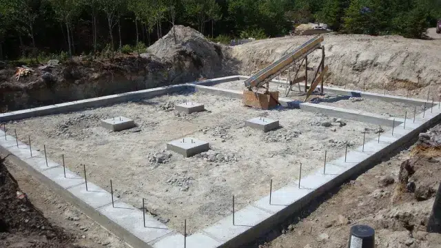 A construction site showing a freshly poured concrete foundation with reinforcing rebar protruding from the slab. The rectangular foundation pad is surrounded by exposed earth and gravel piles, with construction equipment visible in the background. Trees line the perimeter of the cleared construction area.