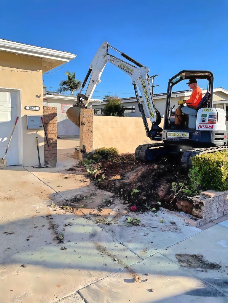 A compact excavator operated by a worker in an orange shirt digs up a front yard area near a residential home's driveway. The excavation has exposed dark soil and damaged concrete pavement, with debris scattered across the ground. The work is taking place in a suburban neighborhood under clear blue skies, with palm trees and houses visible in the background.