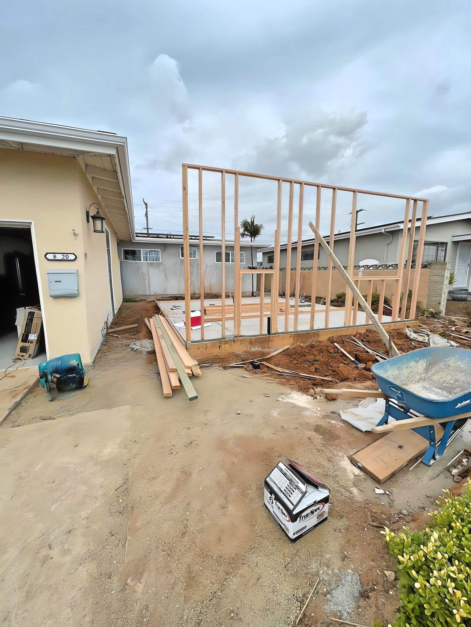 A residential construction site showing the wooden frame structure of a new addition being built next to a pale yellow house. The skeletal frame stands on a concrete foundation surrounded by construction materials including lumber, tools, a blue wheelbarrow with concrete, and disturbed dirt. Dark storm clouds gather overhead in the suburban neighborhood setting.