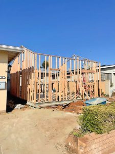 A residential construction site showing the wooden frame structure of a home addition or new build in progress. The skeletal frame made of light-colored lumber stands against a clear blue sky, with an existing single-story residence visible in the background. The construction area includes exposed red dirt, some vegetation along a brick border, and appears to be in an established neighborhood.