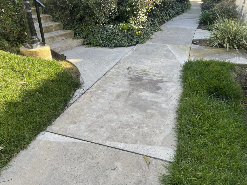 A concrete pathway winds through a landscaped garden area, flanked by well-maintained green lawn on both sides. The walkway shows some weathering and staining on its surface, with visible seams between concrete sections. Stone steps and dense greenery are visible in the background, along with a lamp post on the left side.
