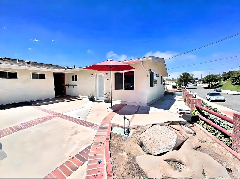 A single-story beige stucco house with a damaged roof sits on a corner lot with multi-level concrete terraces bordered by red brick edging. A red patio umbrella stands near the entrance, and the front yard features multiple levels separated by brick-bordered steps. The property shows signs of needed repairs with exposed dirt patches in the landscaped areas and a residential street visible in the background.