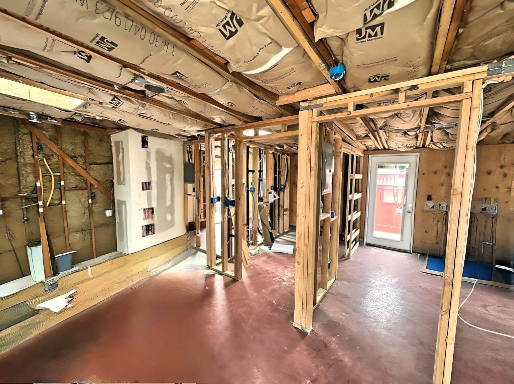 Interior view of a house under renovation showing exposed wooden wall studs and ceiling joists, with visible plumbing and electrical work in progress. The space features a reddish-brown painted concrete floor, insulation materials on the walls and ceiling, and a white exterior door visible in the background. Construction materials and rough framing indicate active remodeling work in what appears to be a basement or lower level.
