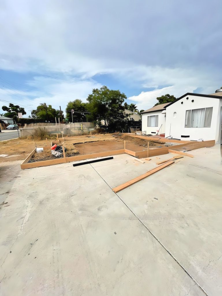 A residential construction site showing wooden frame forms laid out on bare dirt and concrete foundation in preparation for building. A white single-story house with Spanish-style architecture is visible on the right side of the property. The scene includes a cloudy sky, surrounding trees, and utility poles in a suburban neighborhood setting.