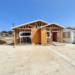 A single-family home under construction showing exposed wooden frame and roof trusses. The structure features plywood sheathing on the walls, window openings, and a concrete foundation and driveway. The construction site is in a residential neighborhood with other houses visible in the background under a clear blue sky.
