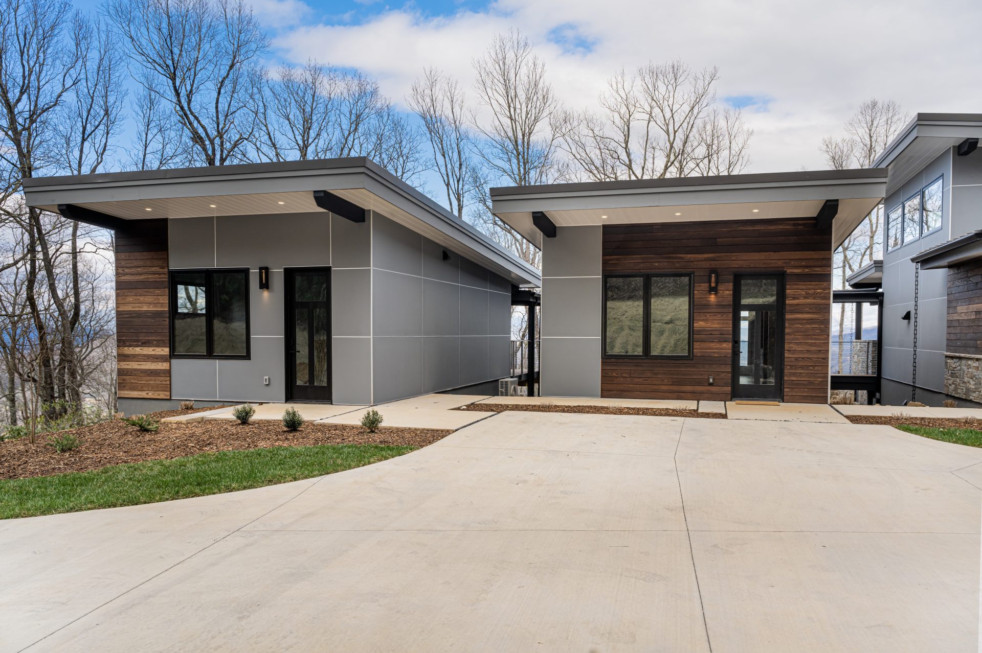 A modern single-story home featuring a contemporary design with clean lines and mixed materials including horizontal wood siding and gray panels. The house has a distinctive angular roofline with wide overhangs, large black-framed windows, and is set on a concrete driveway with landscaped areas of mulch and grass. Bare trees frame the property against a partly cloudy blue sky.