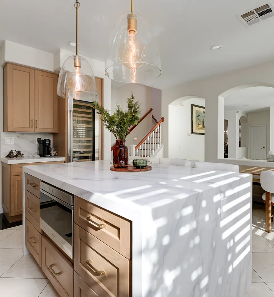 A modern kitchen featuring a large white marble island with natural wood cabinetry and brass hardware. Two contemporary glass pendant lights with copper accents hang above the island, which displays a brown decorative vase with greenery. The bright, open space showcases white walls, an arched doorway, visible staircase, and a wine refrigerator, all bathed in natural sunlight creating geometric shadow patterns on the countertop.