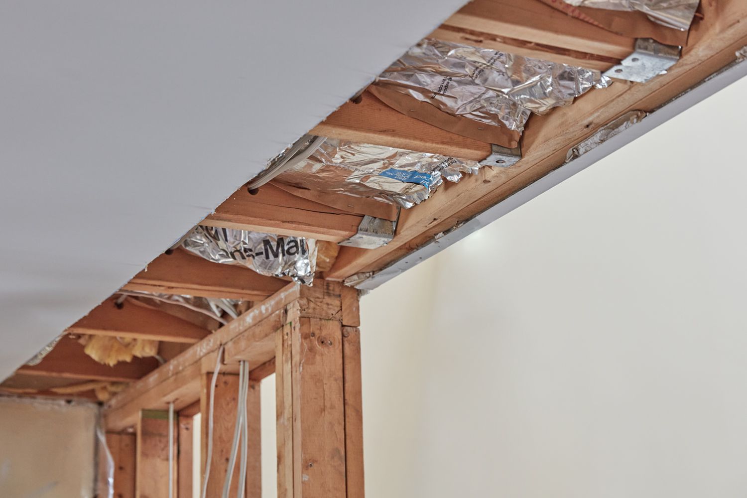 An exposed ceiling and wall corner during home renovation showing wooden joists and studs with reflective silver insulation material installed between the framing members. The drywall has been removed to reveal the interior structure, with white walls visible on the right side and the bare ceiling framing exposed above. The construction scene demonstrates typical residential building materials and insulation installation in progress.