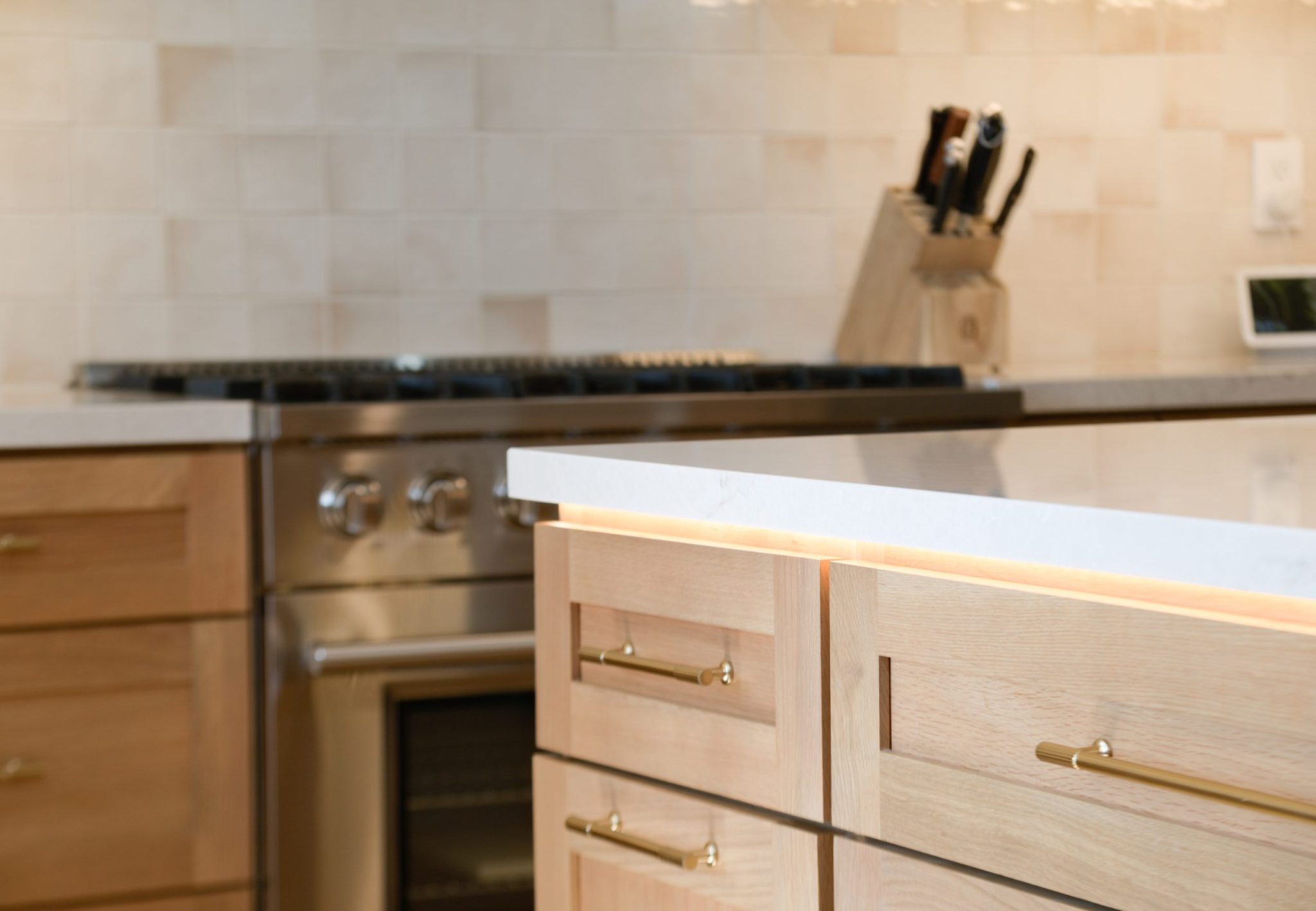 A modern kitchen interior featuring light wood cabinetry with brass pull handles and a white countertop. In the background, a stainless steel range sits beneath a dark countertop, with a wooden knife block visible on the counter. The kitchen has a clean, minimalist design with pale tile backsplash and warm natural wood tones throughout.