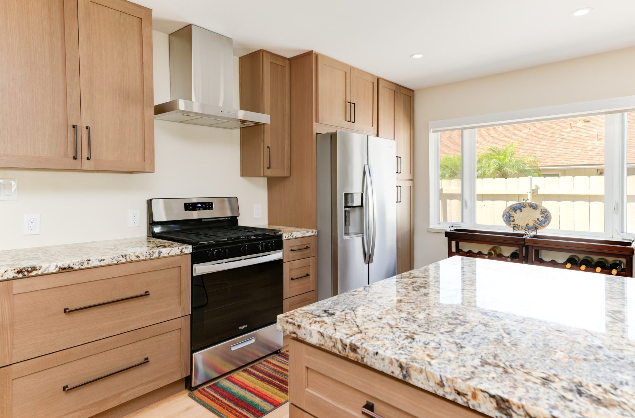 A modern kitchen featuring light wood shaker-style cabinets with dark hardware, complemented by speckled granite countertops. The space includes stainless steel appliances, a gas range with hood, and a central island. Natural light streams through a window overlooking a fenced backyard, with a wooden wine rack visible near the window.