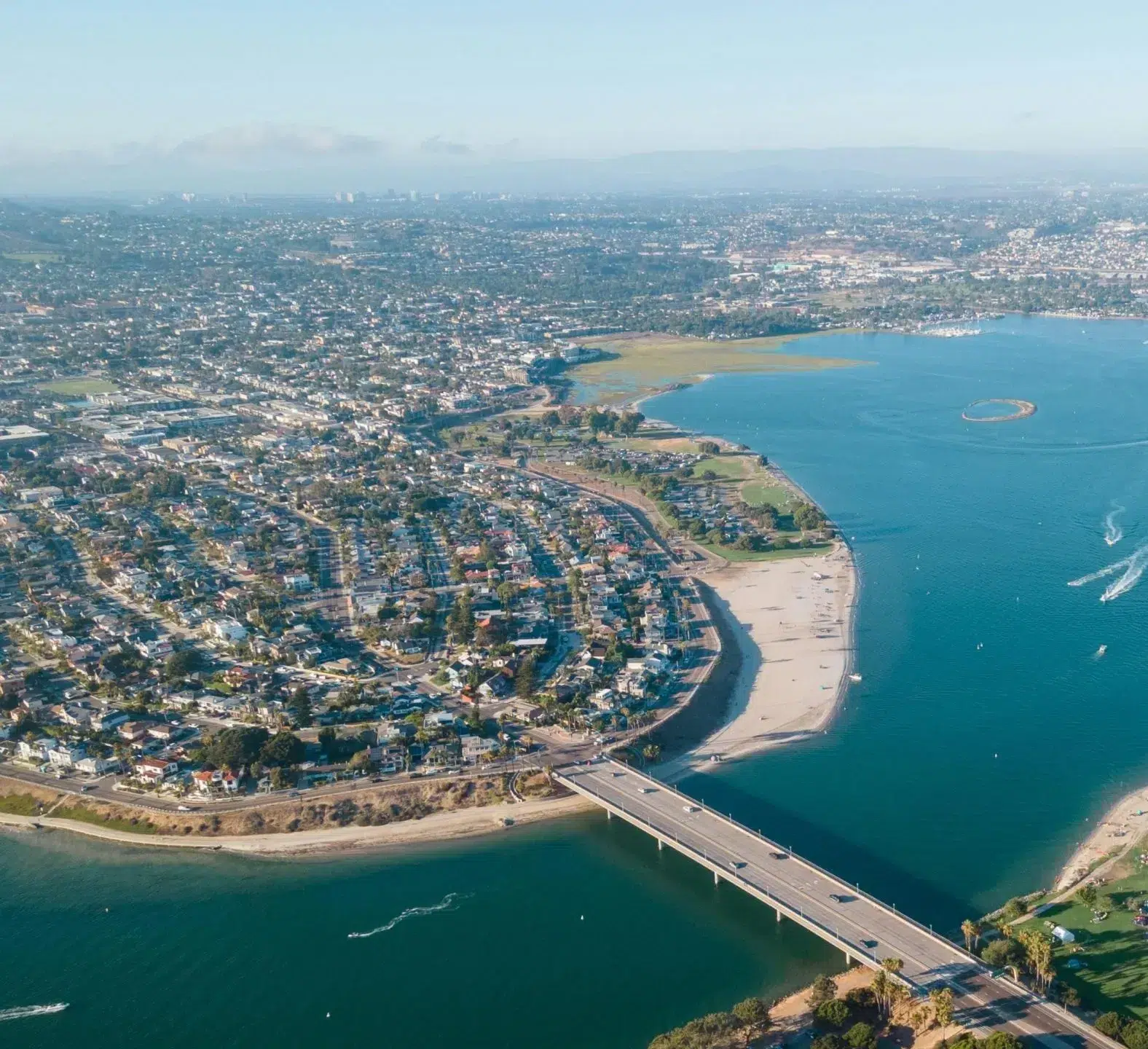 Aerial view of a coastal residential area with a long bridge crossing a turquoise bay or inlet. Dense housing developments line the curved shoreline with sandy beaches visible along the water's edge. The urban landscape extends into the hazy distance with mountains visible on the horizon.