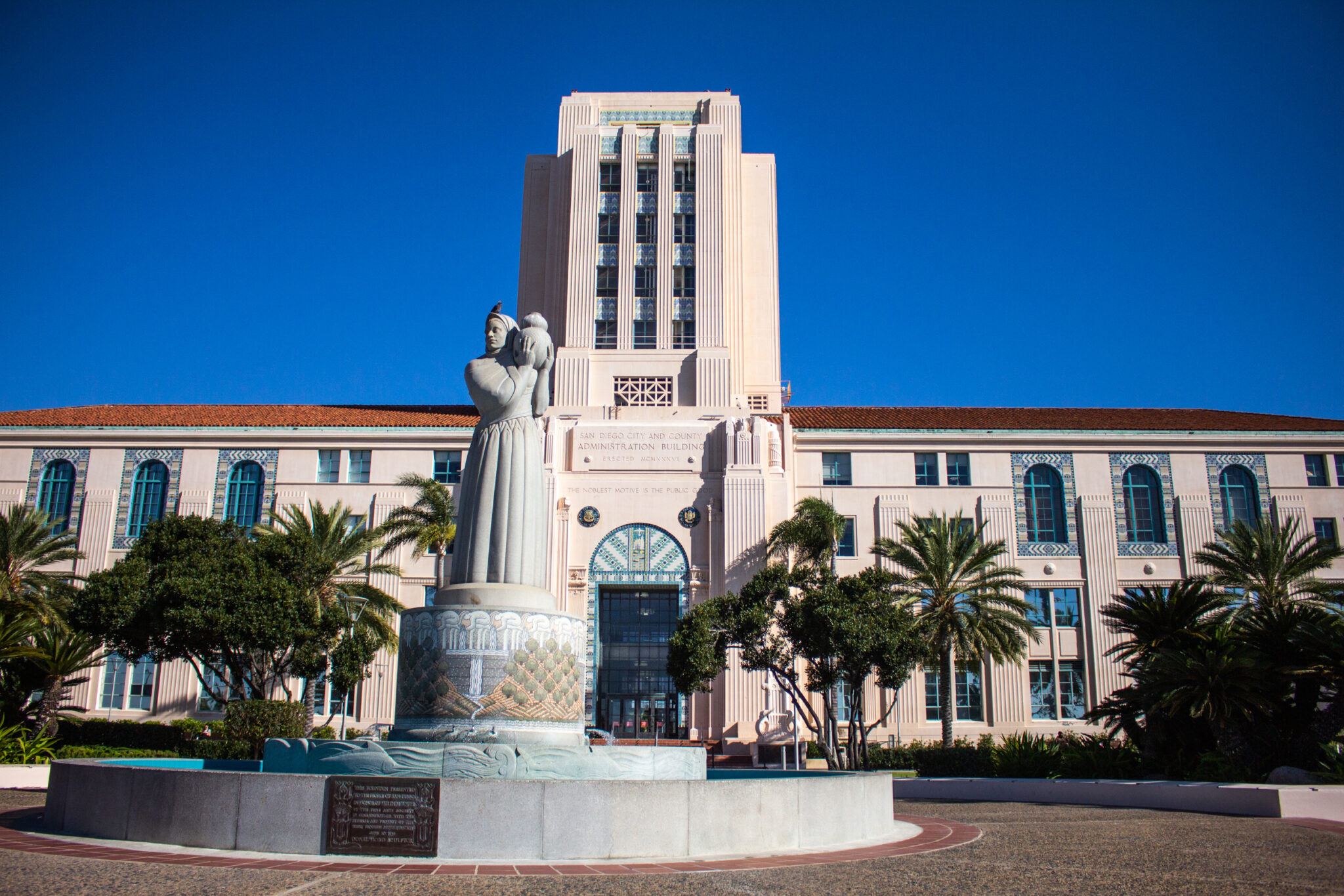 A grand Art Deco government building with a prominent central tower features cream-colored facade and decorative vertical lines. In the foreground, a large white statue monument depicts a standing figure atop an ornamental base. Palm trees and manicured landscaping frame the building's entrance against a brilliant blue sky.