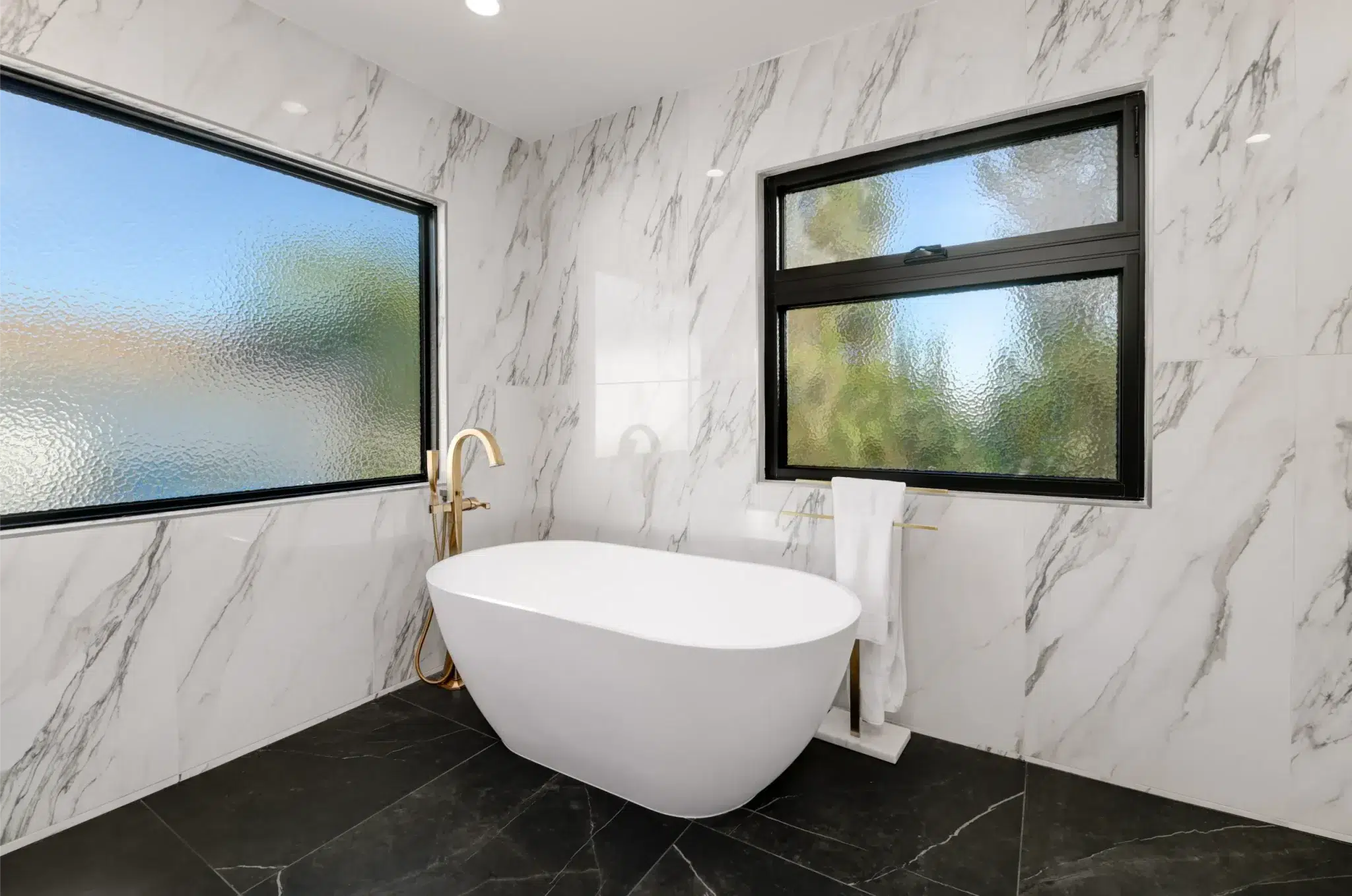 A modern luxury bathroom featuring a white freestanding soaking tub centered in a corner beneath two black-framed windows with frosted glass. The space showcases elegant white marble walls with gray veining and contrasting black marble flooring. Gold fixtures complement the minimalist design, with natural light filtering through the textured glass windows.
