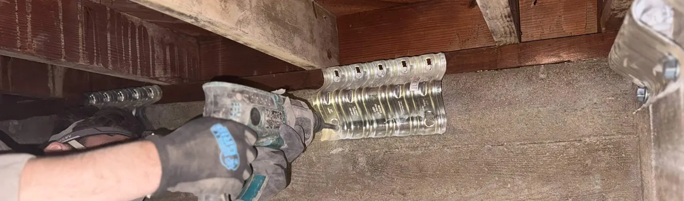 A gloved hand operates a caulking gun to apply sealant along a gap between wooden structural elements and concrete or masonry surfaces in what appears to be a basement or foundation area. The image shows home weatherization or insulation work being performed to seal air leaks. Various wooden framing members and foundation materials are visible in the dusty, unfinished space.