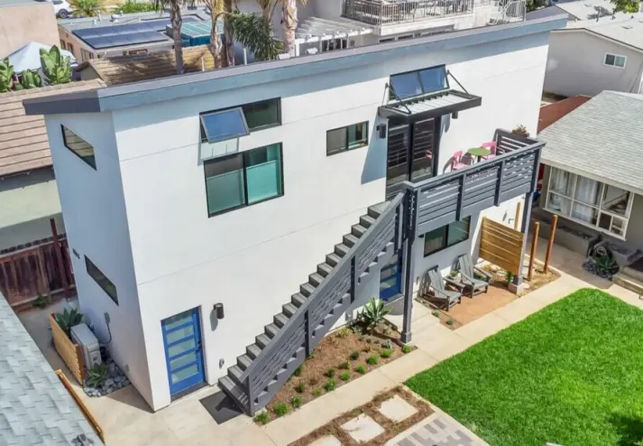 Aerial view of a modern two-story white residential home featuring a flat roof with solar panels, contemporary architectural design with clean lines, and a well-maintained backyard with green lawn, patio furniture, and outdoor living space. The property includes black privacy screening on an exterior staircase and is situated in a suburban neighborhood with adjacent houses visible on either side.
