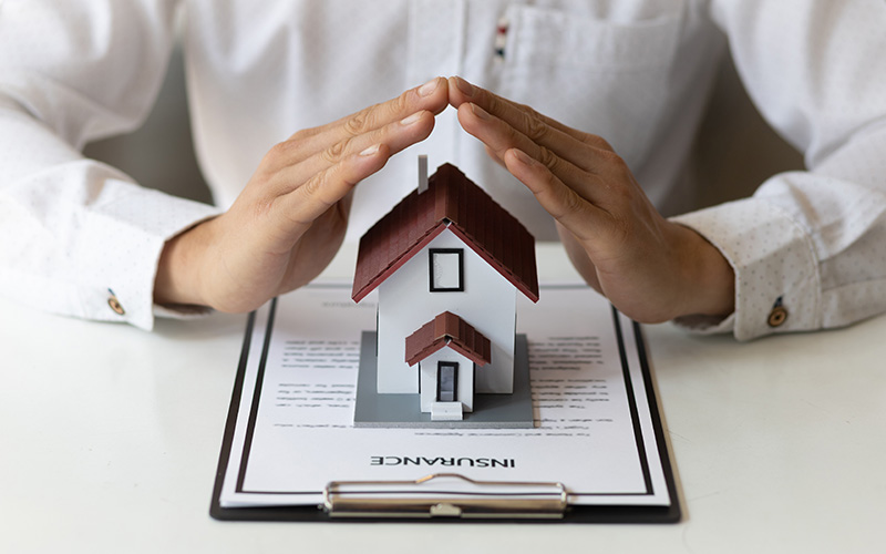 A person in a light-colored business suit holds their hands in a protective arch position over a small model house with a red roof, which sits on top of an insurance document attached to a clipboard. The image symbolizes home insurance protection and property coverage, with the hands representing the security and safeguarding that insurance policies provide to homeowners.