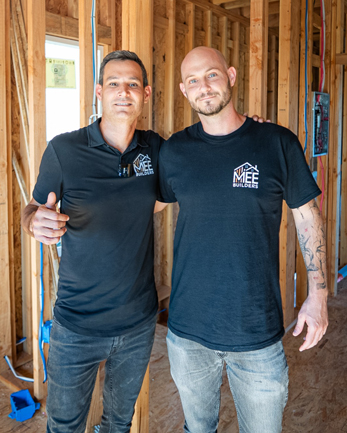 Two men standing side by side in a construction site with exposed wooden framing visible in the background. Both are wearing dark shirts with what appears to be a company logo. The man on the left wears a polo-style shirt while the man on the right wears a t-shirt and has visible tattoos on his arm.