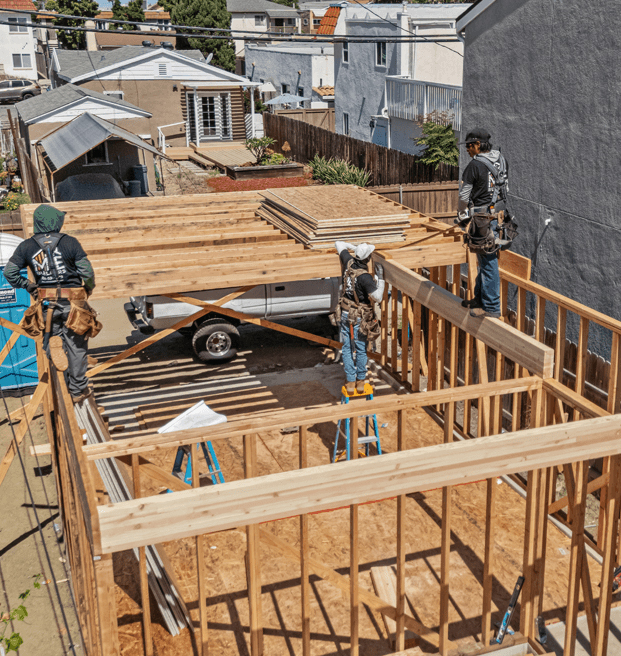 Construction workers build wooden frames for a new residential structure on a rooftop in a densely populated urban neighborhood. The framing shows the skeletal outline of rooms with exposed studs, while plywood sheets are stacked on a trailer below. Surrounding buildings and homes fill the background, illustrating the compact nature of the development area.
