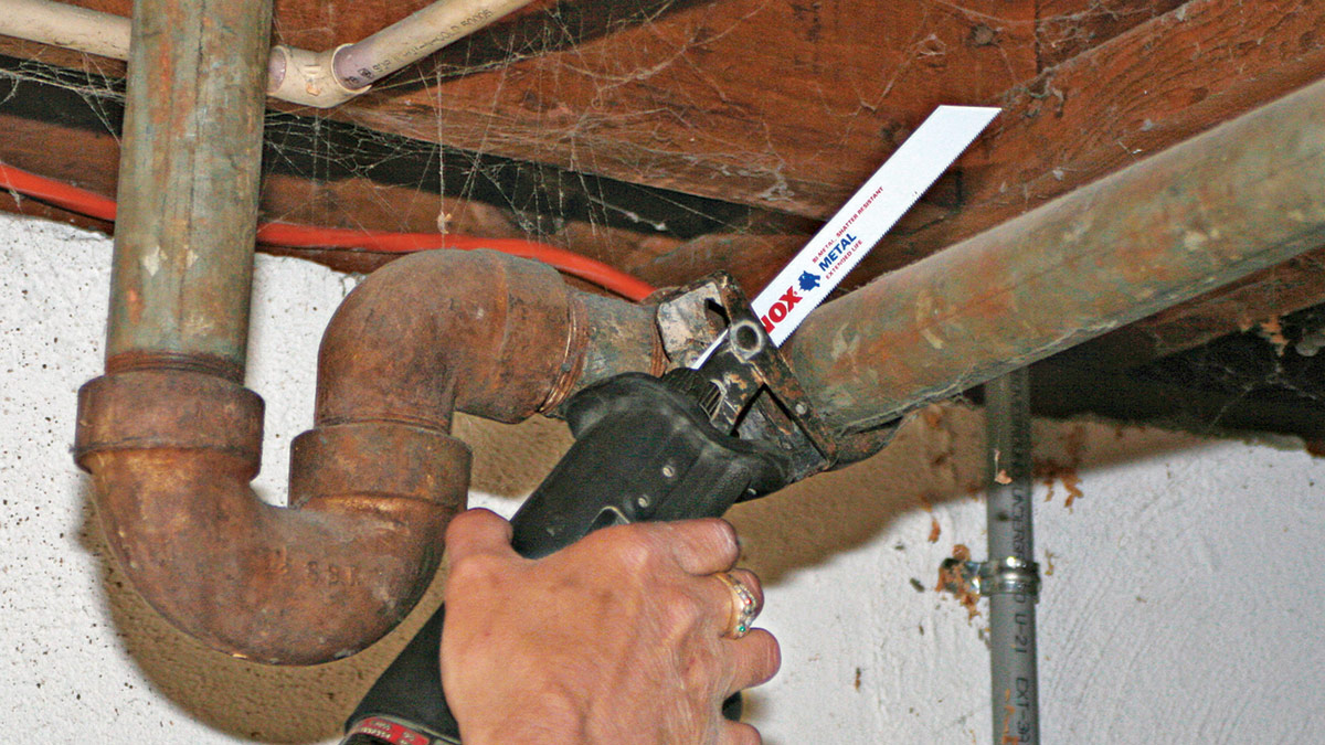 A hand wearing a ring uses a Lenox hacksaw with a metal blade to cut through old, rusted plumbing pipes in what appears to be a basement or crawl space. The workspace shows deteriorated wooden beams covered in cobwebs, corroded metal pipes including a U-shaped trap, and exposed infrastructure. The person is performing plumbing repair or renovation work in confined, aged conditions.
