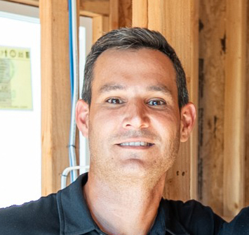 A smiling man with dark hair wearing a black polo shirt poses for a headshot in an indoor setting with wooden framing and windows visible in the background. The photo appears to be a professional or casual portrait taken in what looks like a construction site or building under renovation. The image has a warm, approachable quality with natural lighting from the windows.