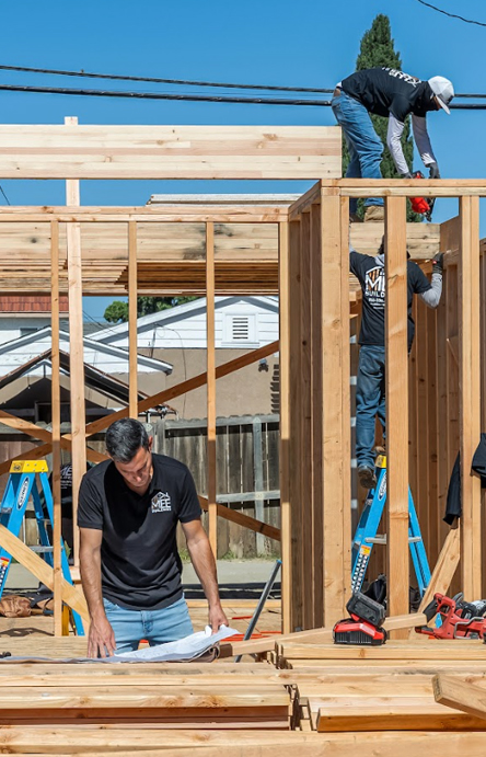Construction workers building a wooden frame structure on a residential job site. The image shows exposed framing with vertical studs and horizontal beams, with workers actively engaged in the building process. Natural wood materials are visible throughout the structure, and the scene captures an active construction phase of what appears to be a home or building project.