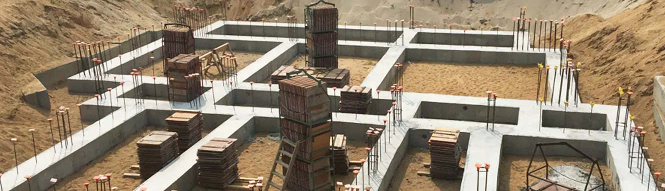 An aerial view of a concrete foundation under construction on a sandy building site. The foundation shows multiple walls and columns with exposed rebar protruding upward, along with wooden formwork and cylindrical column forms. Construction materials and equipment are scattered throughout the work area between the foundation walls.