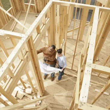 Two construction workers in hard hats review plans or documents while standing inside a wooden frame structure during home construction. The image is taken from an elevated angle, showing exposed wall framing, floor joists, and building materials scattered around the unfinished interior space. The workers appear to be inspecting or discussing the building progress.