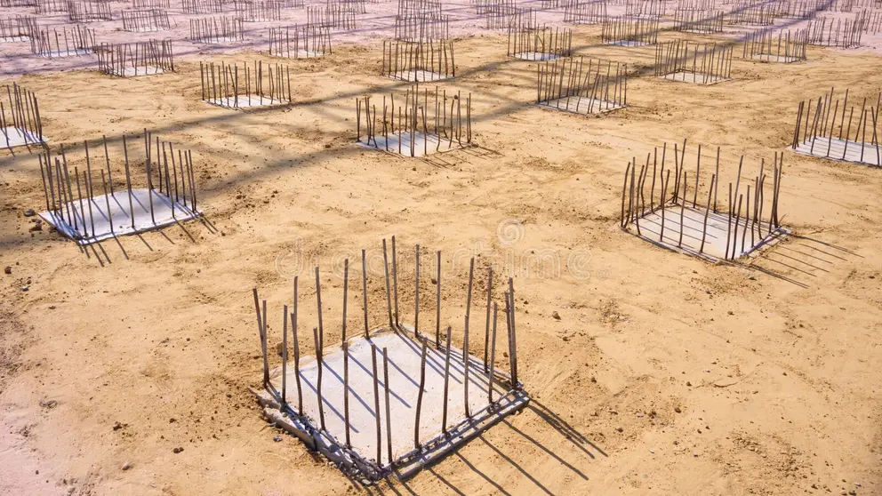 A construction site showing multiple concrete foundation pads with reinforcing steel rebar protruding vertically from each base, arranged in a grid pattern across sandy terrain. The foundations appear to be prepared for column construction, with each pad featuring a systematic arrangement of steel reinforcement bars ready to connect with future structural elements. The image captures an early stage of building construction, likely for a multi-story structure or industrial facility.