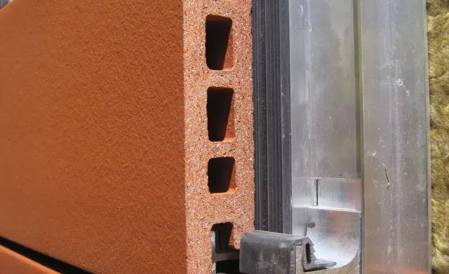 A close-up view of a metal door frame or track system installed against a red-orange brick wall with rectangular hollow openings. The metal frame appears to be aluminum or galvanized steel with mounting hardware visible, likely part of a sliding door or window installation. Yellow insulation material is visible in the background behind the wall assembly.