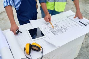 Construction workers reviewing architectural blueprints and floor plans on a concrete surface at a job site. One worker points to details on the plans with a yellow pencil while others examine the documents. A tablet device, yellow hard hat with ear protection, and rolled plans are visible on the work surface.