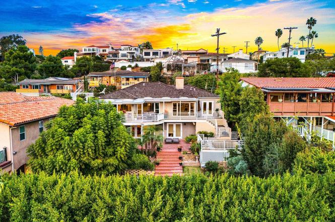A stunning sunset view over a residential neighborhood featuring Mediterranean-style homes with terra cotta tile roofs nestled among lush green vegetation and palm trees. The sky displays vibrant orange, yellow, and blue hues as the sun sets behind the hillside community. Utility poles and a mix of architectural styles, from single-story to multi-level homes with white balconies, characterize this coastal California-style setting.