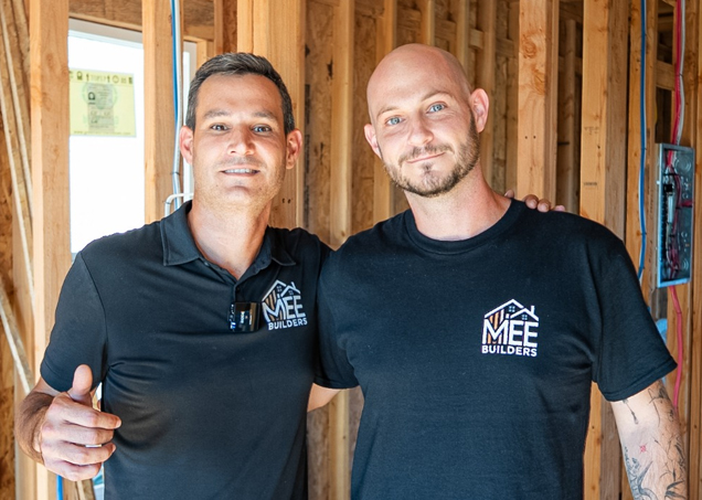 Two smiling construction professionals wearing black MEE Builders branded shirts stand side by side inside an unfinished building with exposed wooden wall framing. The man on the left wears a polo-style shirt while the man on the right wears a t-shirt and has visible tattoos on his arm. Natural light streams through a window opening behind them in the construction site.