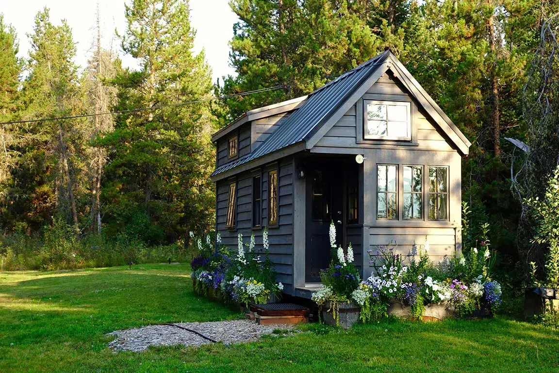 A small dark gray tiny house with a steep pitched metal roof sits on a lush green lawn surrounded by tall evergreen trees. The cottage features large windows, a black front door, and decorative flower boxes with white and purple blooms along its foundation. The compact dwelling showcases modern tiny home architecture with horizontal siding and an upper loft window beneath the peaked roof.
